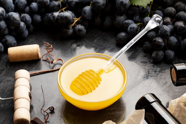 Honey in glass jar with honey spoon on dark stone board with wine cheese corkscrew in Black juicy grapes frame. Delicious yellow bee honey in snack plate on black table.