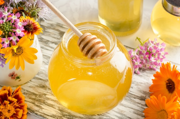 Honey in a glass jar with a honey stick and flowers in a light style