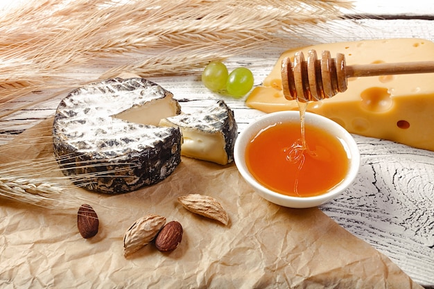 Honey pours into a bowl near cheese, nuts and ears of wheat on crumpled paper and white wooden background.