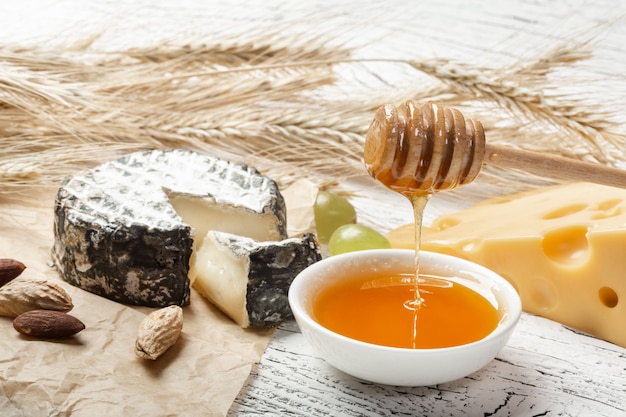 Honey pours into a bowl near cheese, nuts and ears of wheat on crumpled paper