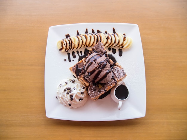 木製のテーブルの上の白い皿にバナナとアイスクリームチョコレートで飾られた蜂蜜トースト