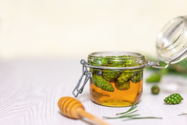 Honey and young green pine cones in an open glass jar on a light wooden table.