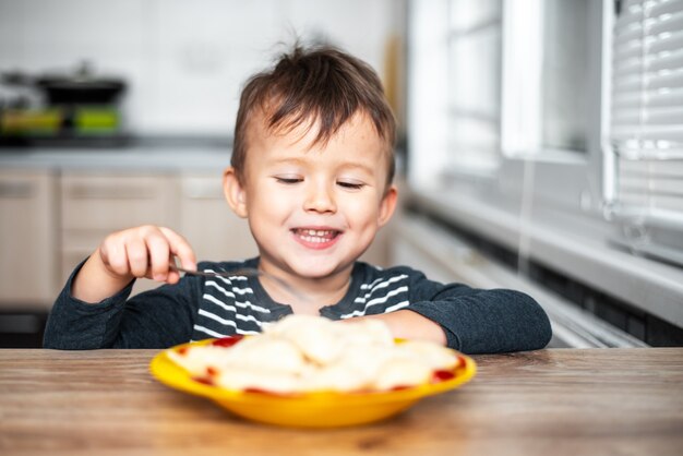 Hongerig kind dat knoedels eet in de keuken, zittend aan tafel in een grijze jas