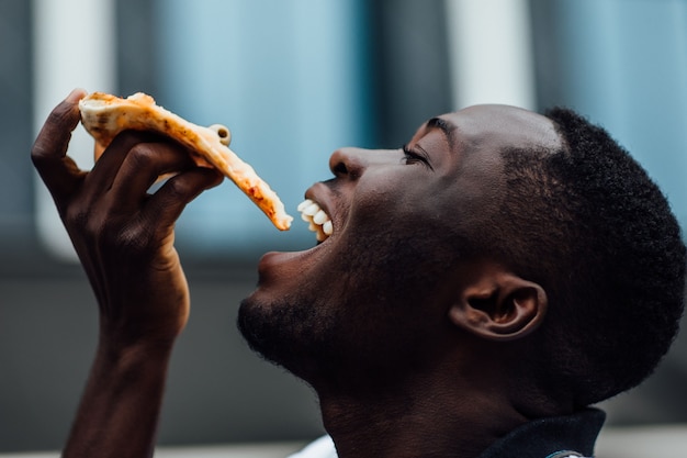 Hongerige afrikaanse man eet pizza op straat close-up portret