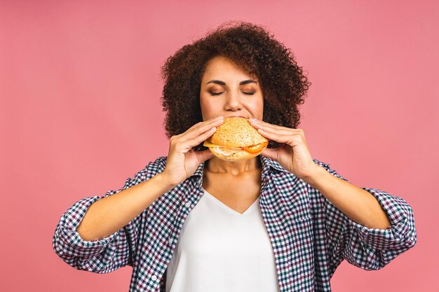 Hongerige Afro-Amerikaanse mooie jonge vrouw die hamburger of burger eet die op roze achtergrond wordt geïsoleerd