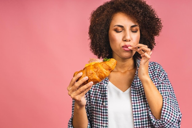 Hongerige Afro-Amerikaanse mooie jonge vrouw eet croissant of sandwich geïsoleerd op roze achtergrond