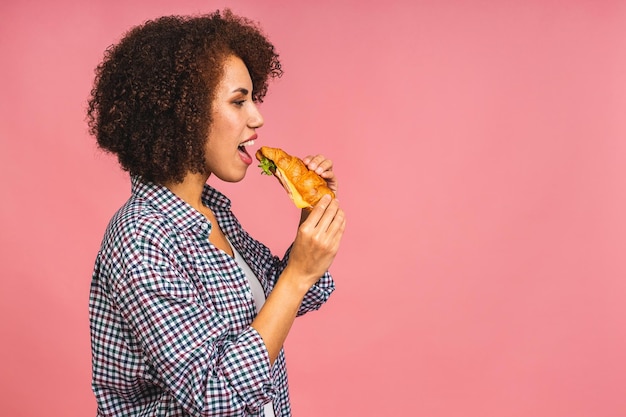 Hongerige Afro-Amerikaanse mooie jonge vrouw eet croissant of sandwich geïsoleerd op roze achtergrond