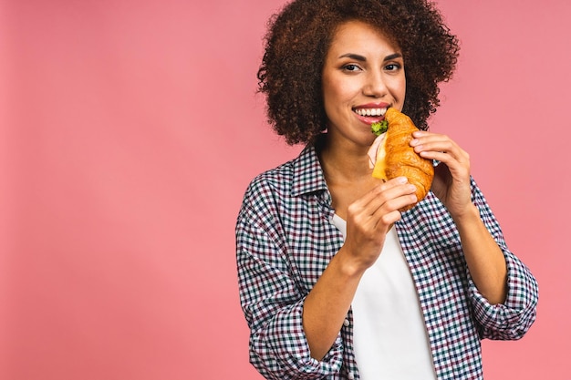 Hongerige Afro-Amerikaanse mooie jonge vrouw eet croissant of sandwich geïsoleerd op roze achtergrond