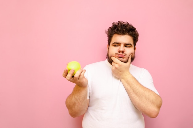 Hongerige dikke man in een wit T-shirt kijkt peinzend naar de appel in de hand