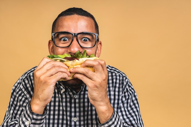 Hongerige jonge Afro-Amerikaanse zwarte man die hamburger eet geïsoleerd over beige achtergrond Dieet of Fastfood concept