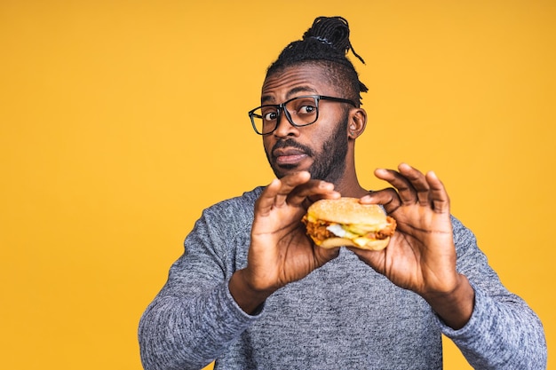 Hongerige jonge Afro-Amerikaanse zwarte man eten hamburger geïsoleerd op gele achtergrond. Dieet concept.