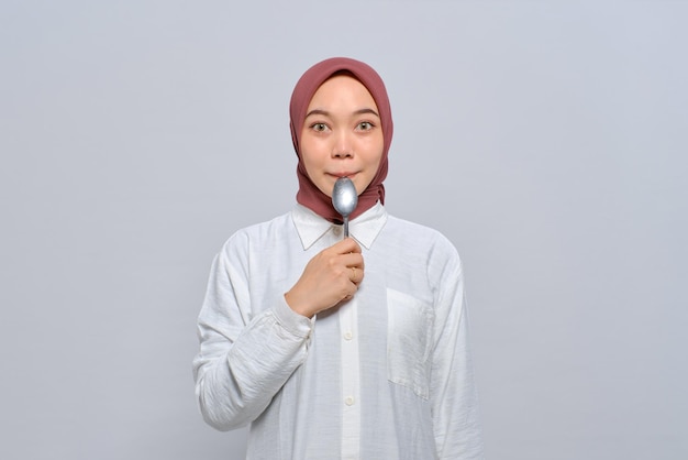 Hongerige jonge Aziatische moslimvrouw met lepel in haar mond dromend over heerlijk gezond voedsel geïsoleerd op witte achtergrond