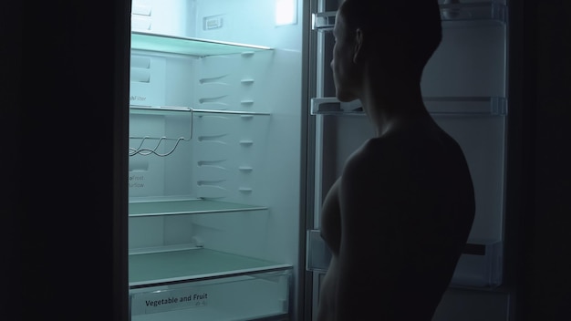 Hongerige man is op zoek naar voedsel om 's nachts in een lege koelkast te eten. Ongezond voedselconcept