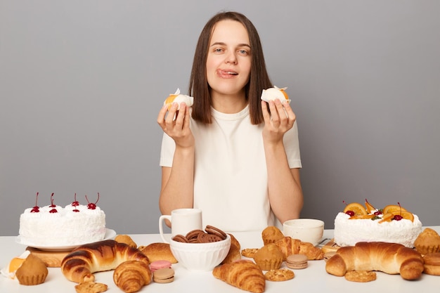 Hongerige mooie vrouw, gekleed in een witte t-shirt geïsoleerd over grijze achtergrond, zittend aan een feestelijke tafel tussen gebak met taarten die hun tong laten zien