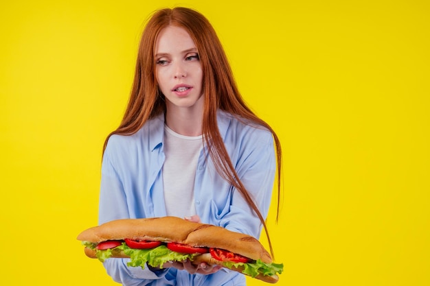 Hongerige roodharige gembervrouw met enorme sandwich met peper en slasalade tussen broodje in studio gele achtergrond.