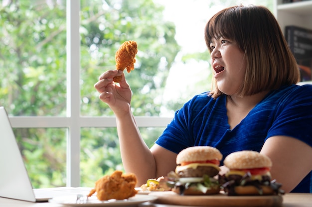 Hongerige vrouw met overgewicht die Fried Chicken, hamburger op een houten bord en pizza op tafel vasthoudt, tijdens het werk vanuit huis, gewichtsprobleem krijgen. Concept van eetbuistoornis (BED).