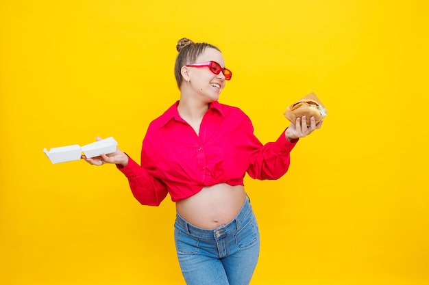 Hongerige zwangere vrouw met hamburger eten van junkfood poseren op gele achtergrond in studio Vrouw genieten van een grote hamburger Het concept van ongezond eten en te veel eten tijdens de zwangerschap