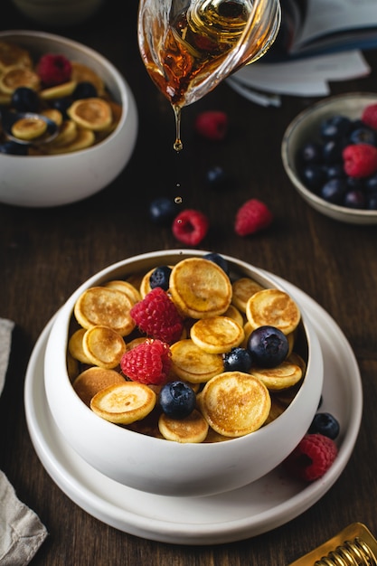 Honing druppels gieten over plaat met pannenkoek granen, trendy mini pannenkoeken met frambozen, bosbessen op houten tafel