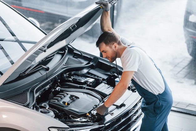 Hood is opened Young man in white shirt and blue uniform repairs automobile