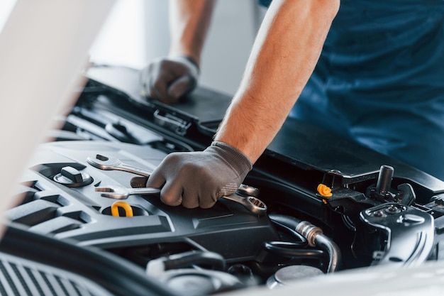 Hood is opened Young man in white shirt and blue uniform repairs automobile