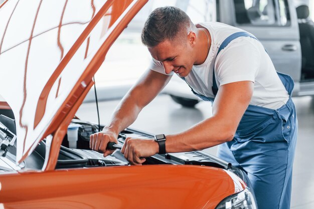 Photo under the hood man in uniform is repairing broken automobile indoors