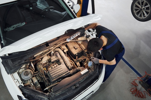 Under the hood Man in work uniform repairs white automobile indoors Conception of automobile service