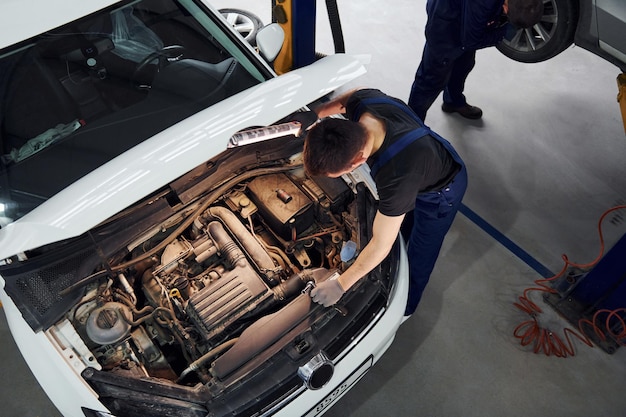 Under the hood man in work uniform repairs white automobile\
indoors conception of automobile service