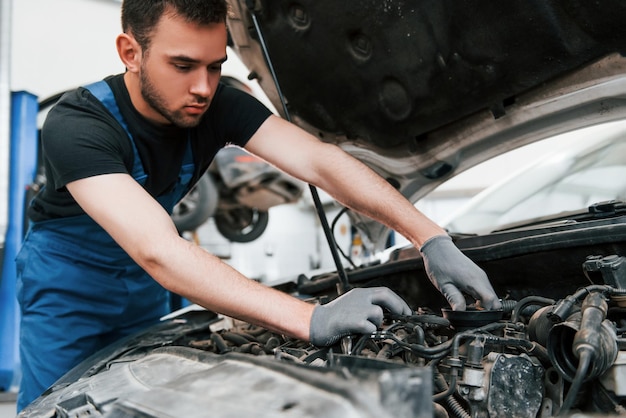 Under the hood man in work uniform repairs white automobile\
indoors conception of automobile service