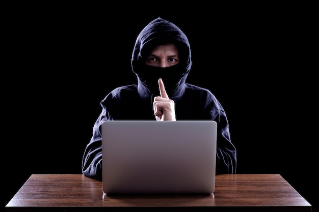 Hooded computer hacker stealing information with laptop Dark background