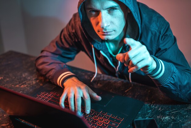 A hooded hacker is typing on a laptop keyboard in a dark room under a neon light