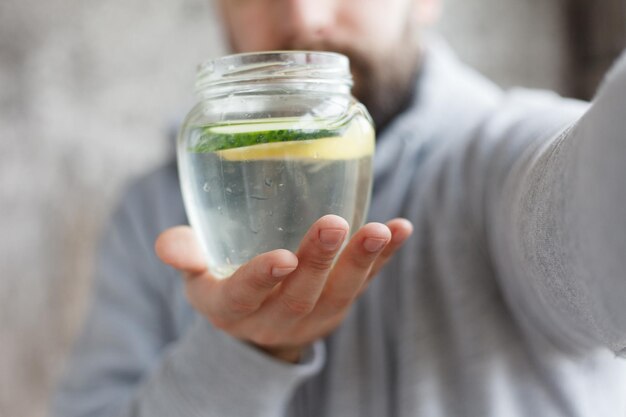 写真 スローモーションで水を飲むフードをかぶった男性グラスにレモンとキュウリのスライスを入れた新鮮な飲み物