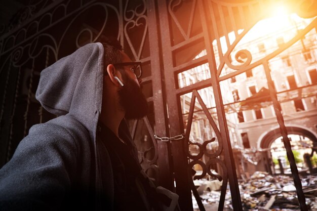 Photo a hooded man with glasses and a beard looks up at a destroyed house in the city center due to the shelling of russian troops