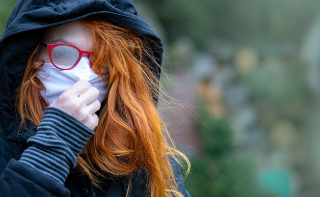 Photo a hooded woman with red hair stands lonely and sad in a park she wears a breathing mask