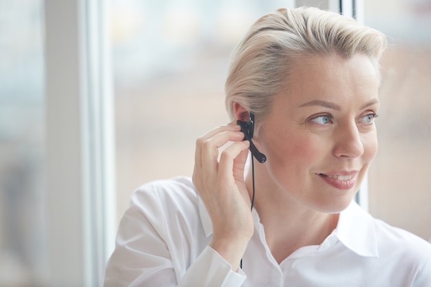 Hoofd en schouders portret van glimlachende zakenvrouw hoofdtelefoon dragen en raam kijken tijdens het werken in callcenter