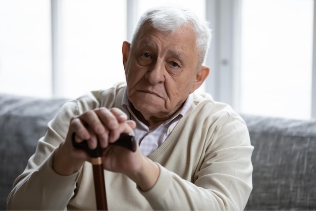 Hoofd geschoten portret depressief eenzame oudere man met houten stok kijkend naar camera gefrustreerd volwassen man gevouwen handen op wandelstok zittend op de Bank alleen eenzaamheid en eenzaamheid