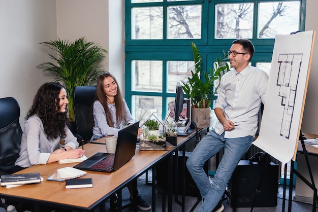 Hoofdingenieur die over nieuw creatief idee in de bouwplannen spreekt terwijl twee vrouwelijke medewerker die in het notitieboekje in het moderne bureau opmerken.