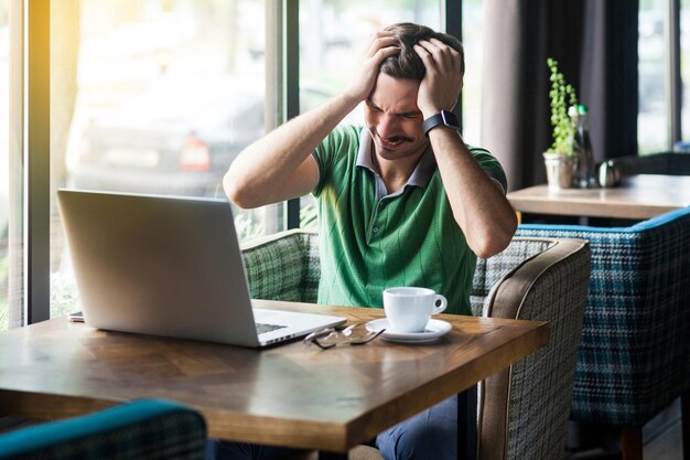 Hoofdpijn probleem of verwarring Jonge zakenman in groene tshirt zitten en houden zijn pijnlijke hoofd zakelijke en freelancen concept binnen schot in de buurt van groot raam overdag