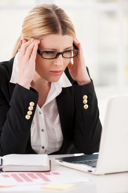 Hoofdpijn voelen. Depressieve volwassen vrouw in formalwear die het hoofd in handen houdt en naar de laptop kijkt terwijl ze op haar werkplek zit