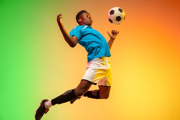 Hoog springen. Mannelijke voetbal, voetballer training in actie geïsoleerd op gradiënt studio achtergrond in neonlicht. Concept van beweging, actie, prestaties, gezonde levensstijl. Jeugd Cultuur.