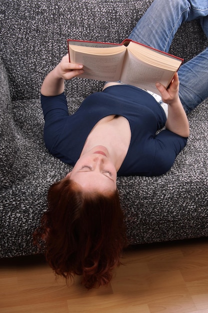Hooghoekbeeld van een jonge vrouw die een boek leest terwijl ze thuis op de bank ligt