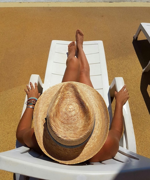 Hooghoekbeeld van een jonge vrouw met een hoed die tijdens een zonnige dag op een stoel op het strand ontspant