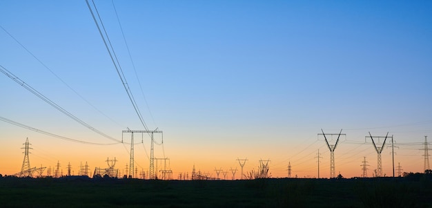 Hoogspanning elektriciteitstorens op veld bij zonsondergang en blauwe lucht panoramisch zicht van donkere silhouetten van hoogspanningslijnen op oranje zonsopgang elektriciteitsopwekking transmissie- en distributienetwerk