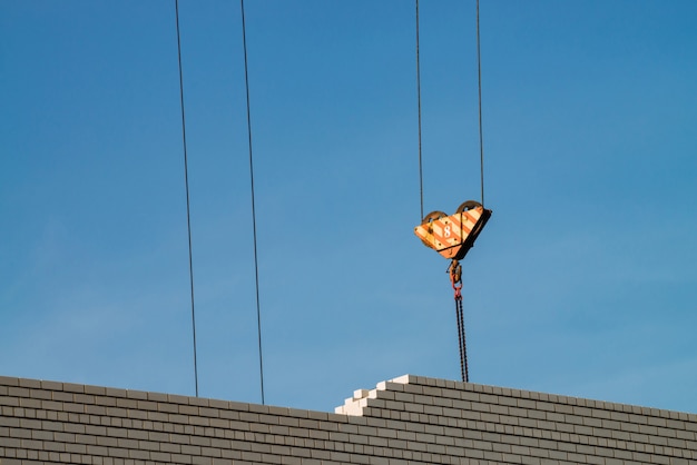 Hook of tower crane above unfinished building in construction site. Image of white brick wall under blue sky.