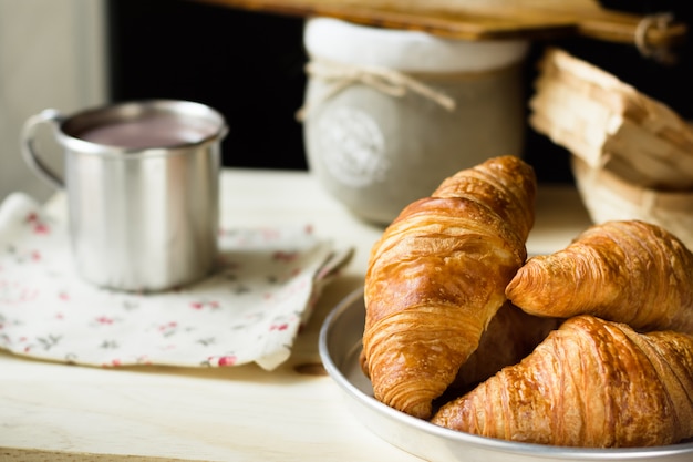 Hoop verse croissants met gouden korst, mok met hete cacao of chocolade, rustieke houten keukentafel