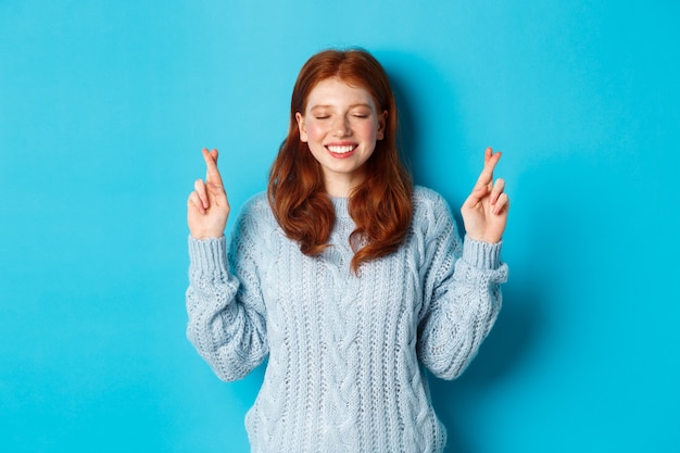 Hoopvol roodharig meisje dat een wens doet, duimen voor geluk, glimlachend en anticiperend op goed nieuws of positief resultaat, zich tegen blauwe achtergrond bevindt.