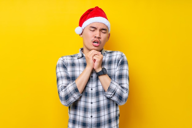 Hoopvolle jonge Aziatische man met een geruit overhemd in een kerstmuts hand in hand gevouwen in gebedsgebaar bedelen om iets op gele achtergrond viering kerstvakantie en nieuwjaarsconcept