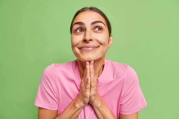 Hoopvolle jonge vrouw houdt handpalmen bij elkaar denkt aan dromen die uitkomen gelooft in een betere glimlach heeft gelukkig donker gekamd haar draagt casual roze t-shirt geïsoleerd over groene muur