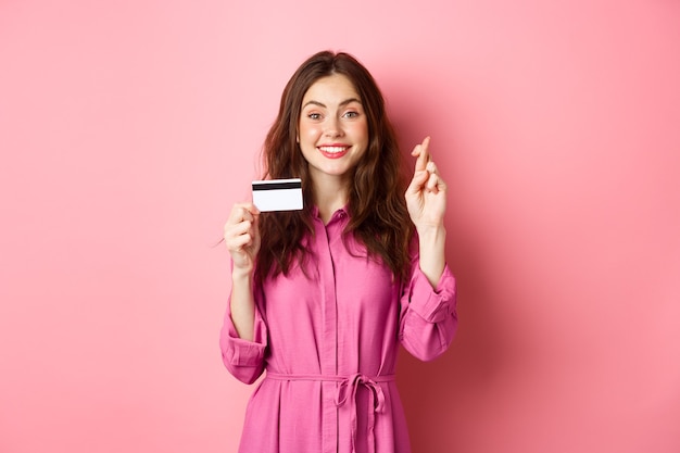 hoopvolle jonge vrouw met plastic creditcard en gekruiste vingers voor geluk, wens doen en glimlachen, staande tegen roze muur.