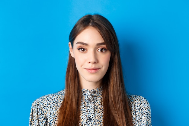 Hoopvolle spontane vrouw met lang haar en make-up, camera kijken met glimlach, staande tegen blauw.