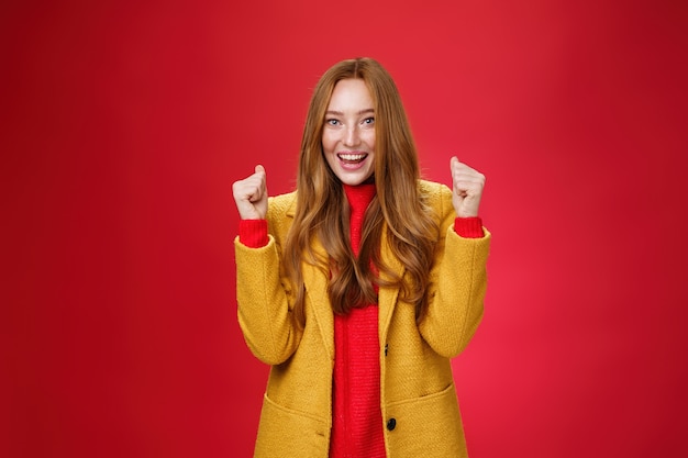 Hooray success portrait of happy cheerful and sincere cute ginger woman in yellow coat raising clenc...